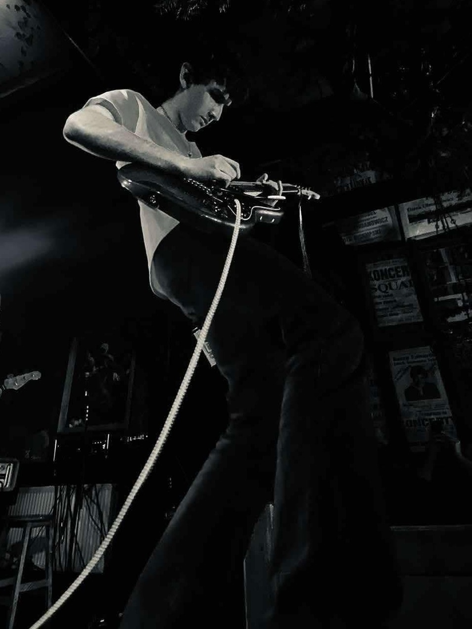 Czarno-białe zdjęcie muzyka grającego na gitarze elektrycznej na scenie, z plakatami koncertowymi w tle i widocznym kablem instrumentu.