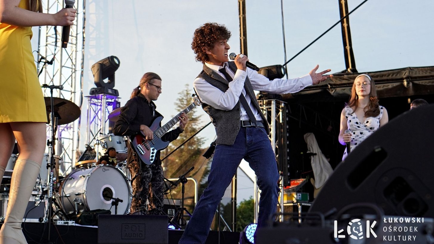 Wokalista z kręconymi włosami śpiewa na scenie z zespołem podczas koncertu plenerowego. Widoczni gitarzysta, perkusja i wokalistki w tle. Logo ŁOK w prawym dolnym rogu.