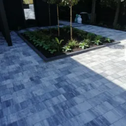 Ułożona szara kostka brukowa z prostokątną rabatą z niską roślinnością i trzema młodymi drzewkami na tle nowoczesnego domu z czarną elewacją.