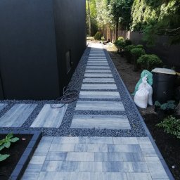 Miki -Bruk - Ułożony chodnik z kostki brukowej w ogrodzie, z ozdobnym żwirem i roślinnością. Widoczny wąż ogrodowy i worki z materiałami budowlanymi. Nowoczesne wykończenie.