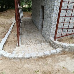 Miki -Bruk - Granitowa ścieżka z obrzeżami prowadzi do kamiennej elewacji budynku. Otwarta, metalowa brama z boku. Widoczne ślady prac ziemnych i fragmenty zieleni w tle.