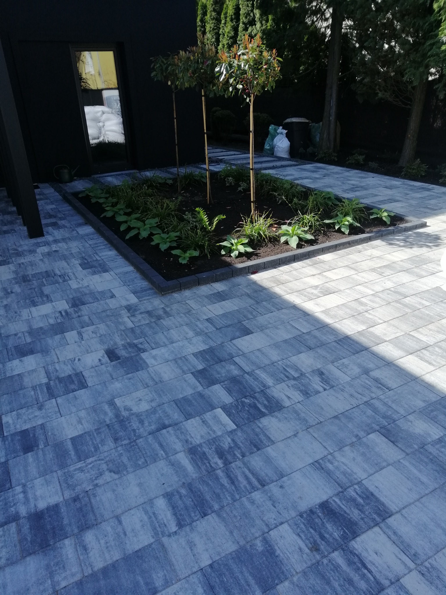 Ułożona szara kostka brukowa z prostokątną rabatą z niską roślinnością i trzema młodymi drzewkami na tle nowoczesnego domu z czarną elewacją.