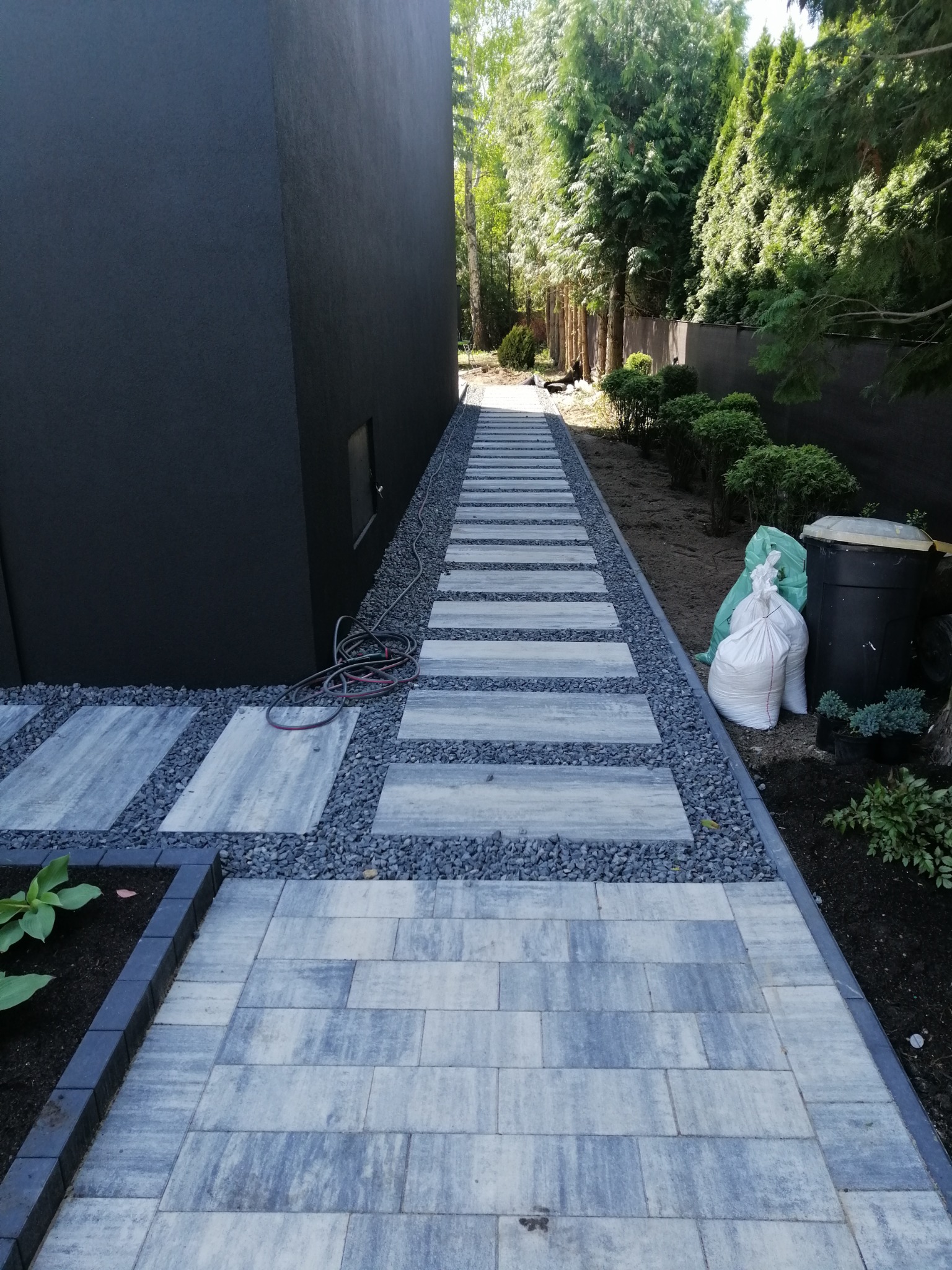 Ułożony chodnik z kostki brukowej w ogrodzie, z ozdobnym żwirem i roślinnością. Widoczny wąż ogrodowy i worki z materiałami budowlanymi. Nowoczesne wykończenie.