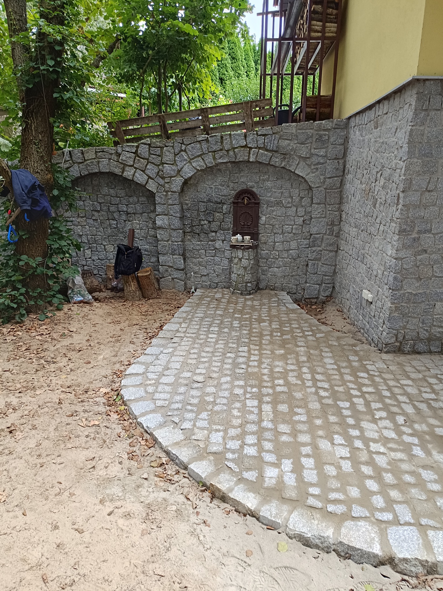 Świeżo ułożona, jasnoszara kostka granitowa tworzy ścieżkę i plac przed kamienną ścianą z dwoma łukami. Na ścianie ozdobna, metalowa dekoracja. Praca w toku, widoczne narzędzia.