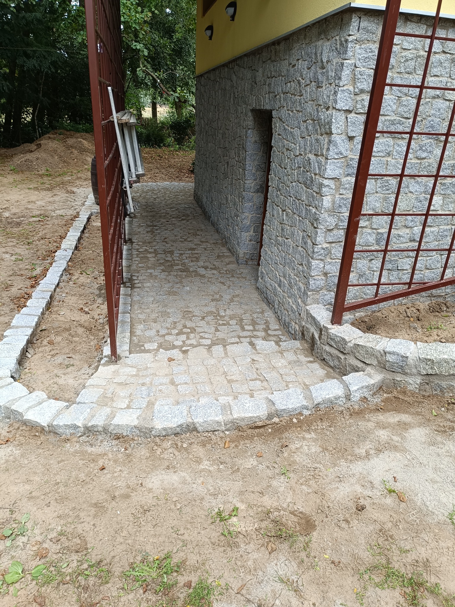 Granitowa ścieżka z obrzeżami prowadzi do kamiennej elewacji budynku. Otwarta, metalowa brama z boku. Widoczne ślady prac ziemnych i fragmenty zieleni w tle.