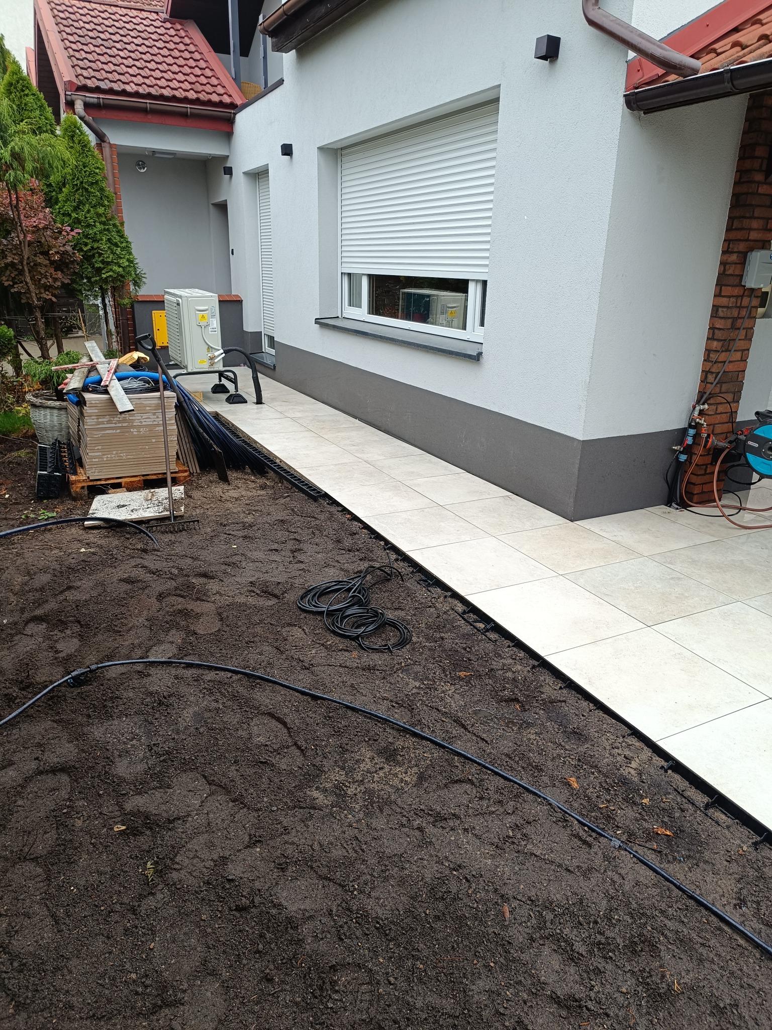 Jasne płytki chodnikowe ułożone wzdłuż ściany domu, kontrastujące z ciemną ziemią i elementami instalacji nawadniającej. Widoczne narzędzia i materiały budowlane.