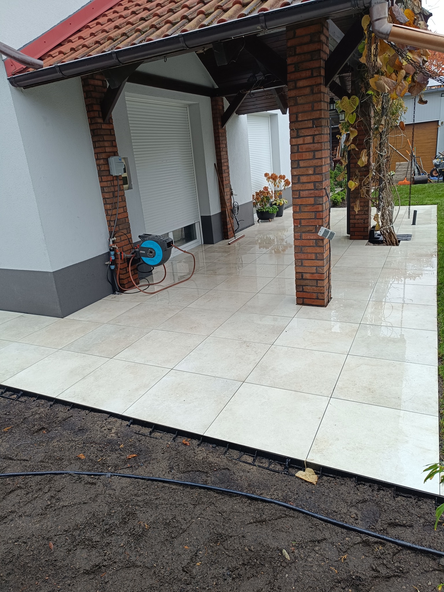 Jasne płytki tarasowe ułożone przy domu z ceglanymi kolumnami i elementami drewnianymi. Widoczny fragment ogrodu i system nawadniania.