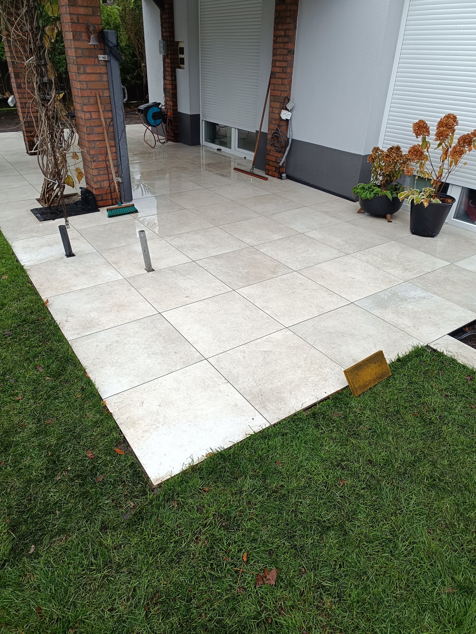 Jasne płytki tarasowe ułożone na zewnątrz domu, z widocznymi elementami trawnika i roślin w donicach. Widoczne narzędzia sugerują prace wykończeniowe.