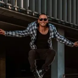 Mężczyzna w okularach przeciwsłonecznych w skoku, ubrany w czarną koszulkę i koszulę w kratę, na tle nowoczesnej architektury.
