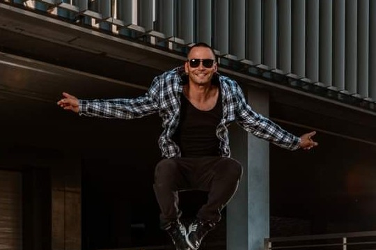 Mężczyzna w okularach przeciwsłonecznych w skoku, ubrany w czarną koszulkę i koszulę w kratę, na tle nowoczesnej architektury.