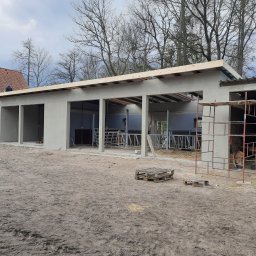 Andrzej Górski, Usługi instalacyjno- budowlane - Surowa elewacja budynku gospodarczego w trakcie budowy, widoczne rusztowania, palety i częściowo wykończony dach, na dachu robotnik.