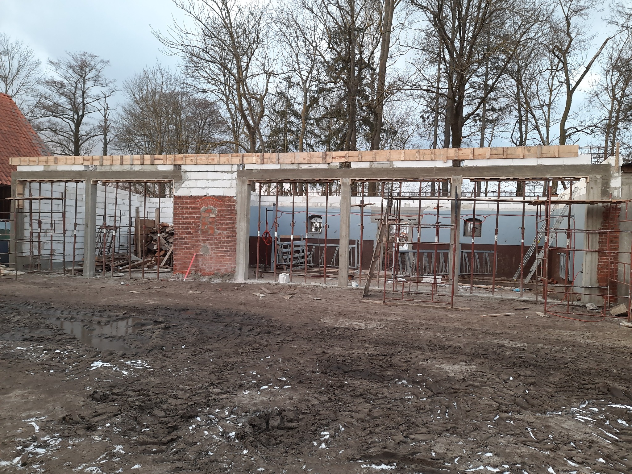 Wznoszona konstrukcja budynku z białych bloczków i czerwonej cegły, widoczne drewniane elementy szalunku i rusztowania na błotnistym terenie.