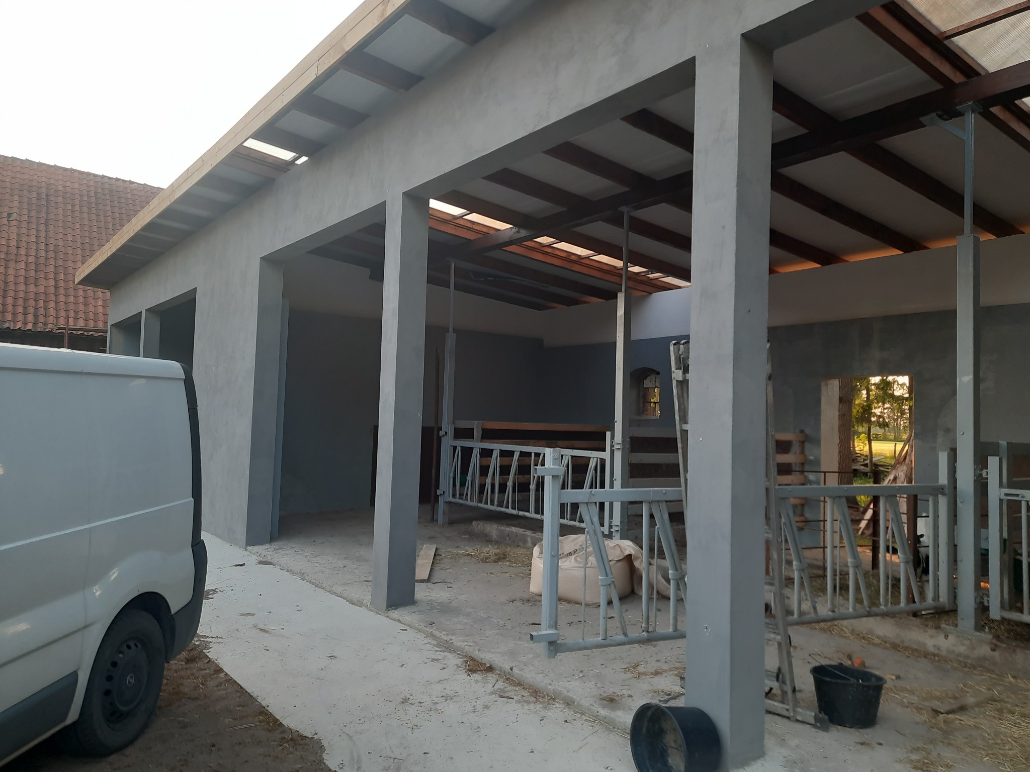 Surowy, szary budynek gospodarczy z częściowo zadaszonymi stanowiskami, widoczny z boku białego samochodu dostawczego, z workami i wiadrami wewnątrz.