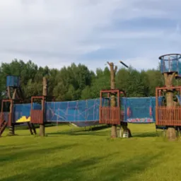 Rozległy park linowy z drewnianymi platformami osadzonymi na drzewach, połączonymi siatkami i mostami linowymi, na tle zielonego trawnika i lasu.