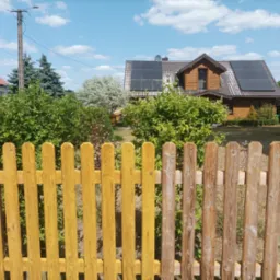 Drewniany dom z panelami słonecznymi na dachu, widoczny zza częściowo pomalowanego na żółto drewnianego płotu.