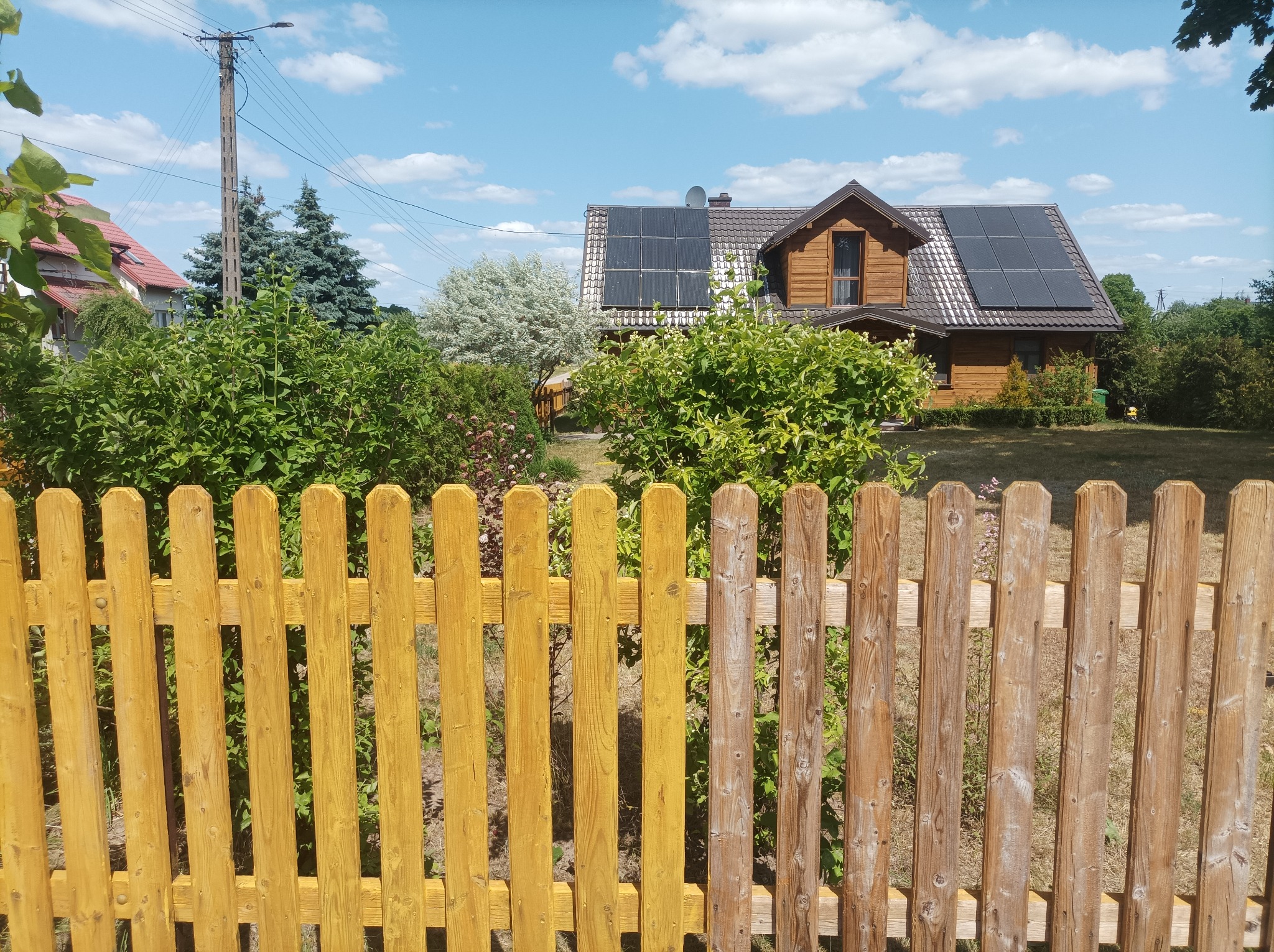 Drewniany dom z panelami słonecznymi na dachu, widoczny zza częściowo pomalowanego na żółto drewnianego płotu.