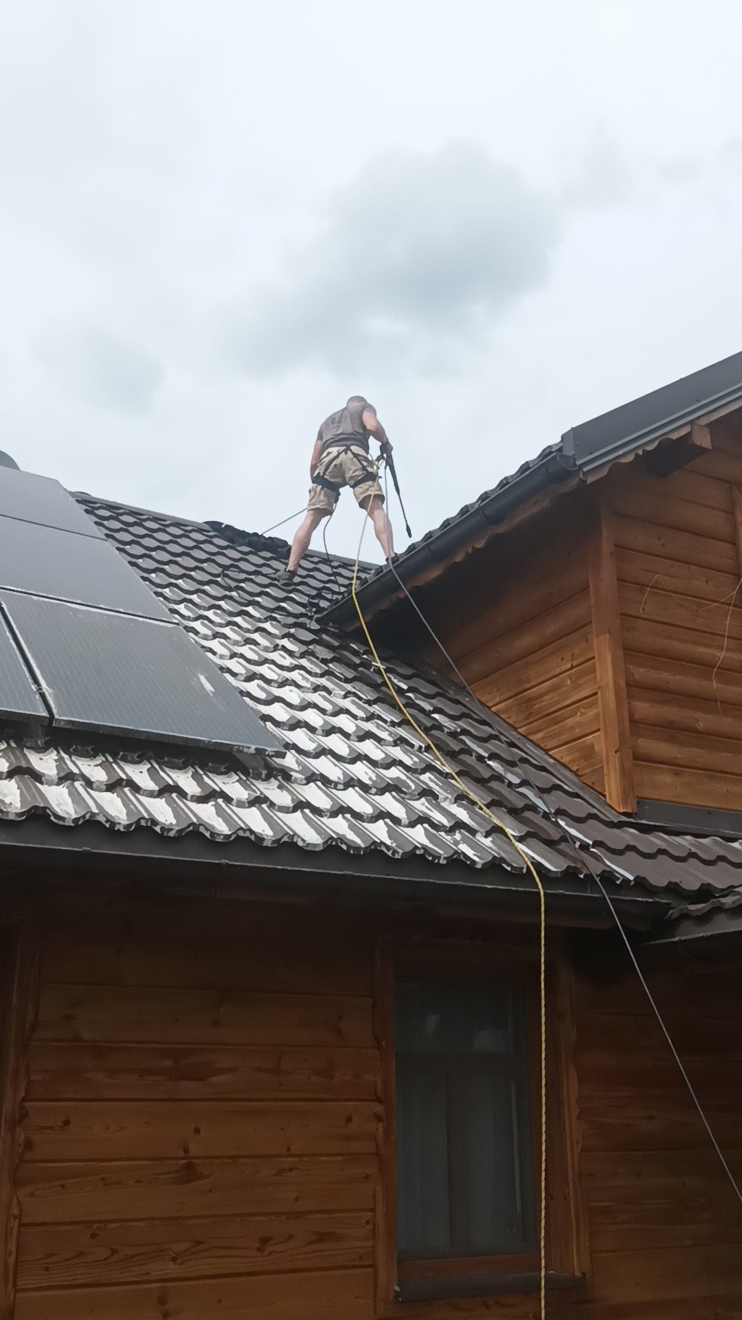 Pracownik w uprzęży zabezpieczającej myje dach ciśnieniowo, w tle brązowa drewniana elewacja domu i panele słoneczne.