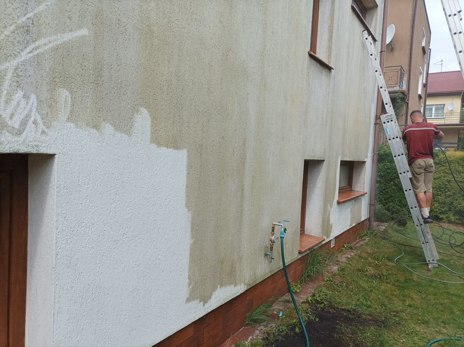 Mężczyzna na drabinie czyści elewację domu myjką ciśnieniową, widoczny fragment umytej i nieumytej ściany, z zielonym wężem ogrodowym i kranem na pierwszym planie.