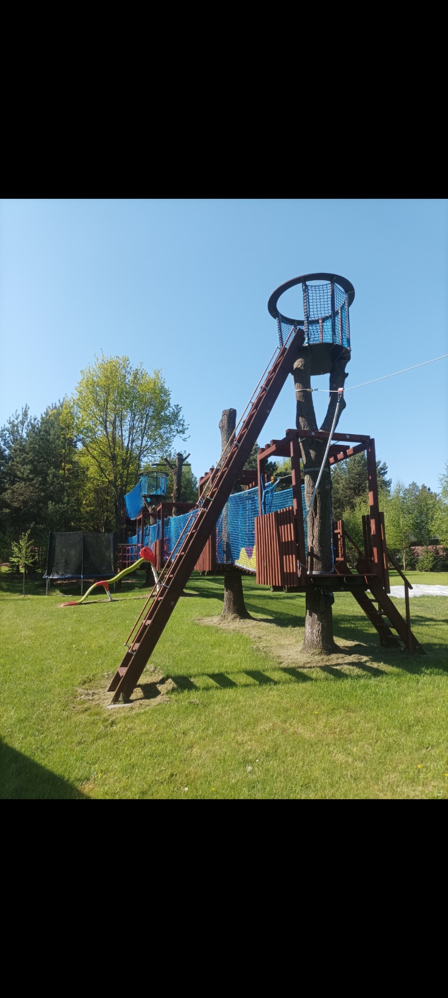 Rozbudowany, drewniany plac zabaw z wieżami, mostami linowymi i zjeżdżalnią, umieszczony na zadbanym trawniku w otoczeniu drzew.