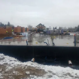 Zalana płyta fundamentowa w trakcie budowy domu jednorodzinnego, widoczne wystające pręty zbrojeniowe, rury instalacyjne, w tle częściowo wymurowane ściany z cegły, koparka i materiały budowlane...