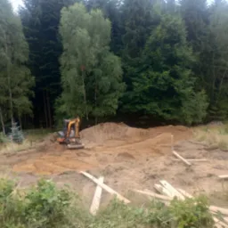 Mała koparka wyrównuje teren pod budowę w otoczeniu drzew, widoczne drewniane elementy szalunku.