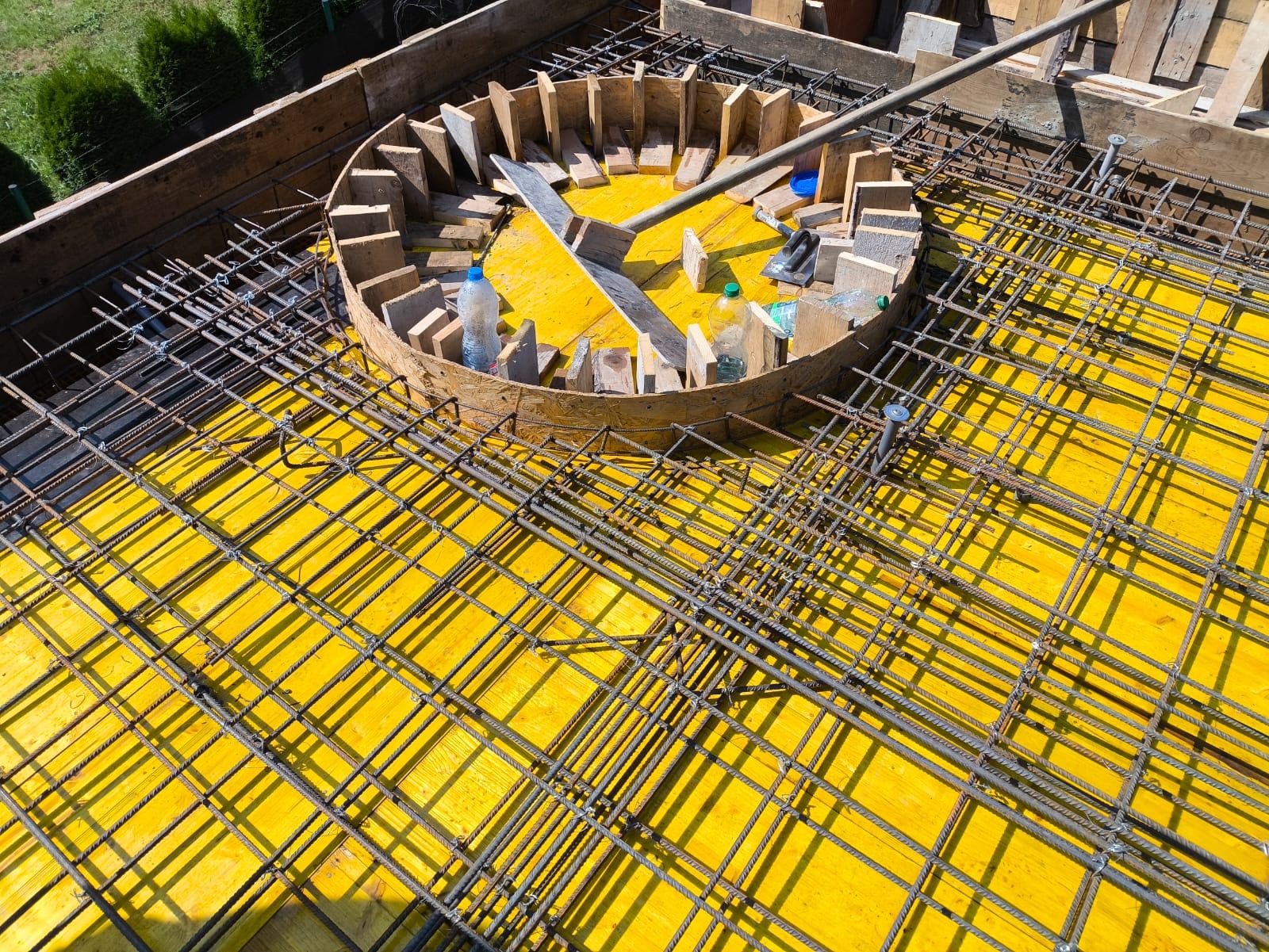 Siatka zbrojeniowa na żółtym szalunku z okrągłym elementem z drewnianych desek, widoczne butelki z wodą i narzędzia, przygotowanie do wylewki betonowej.
