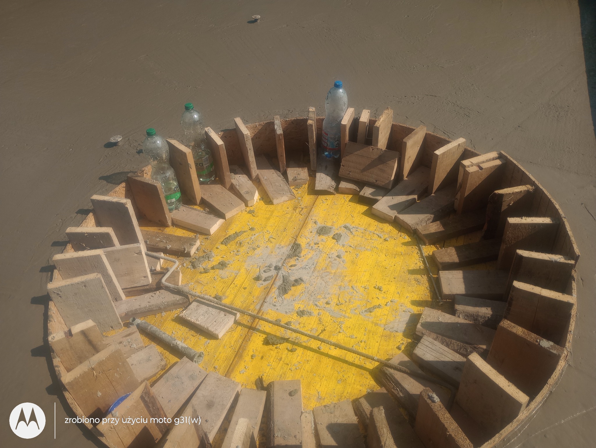 Okrągły szalunek z drewnianych desek na żółtej płycie, zalewany betonem. Widoczne butelki z wodą i pręt zbrojeniowy.