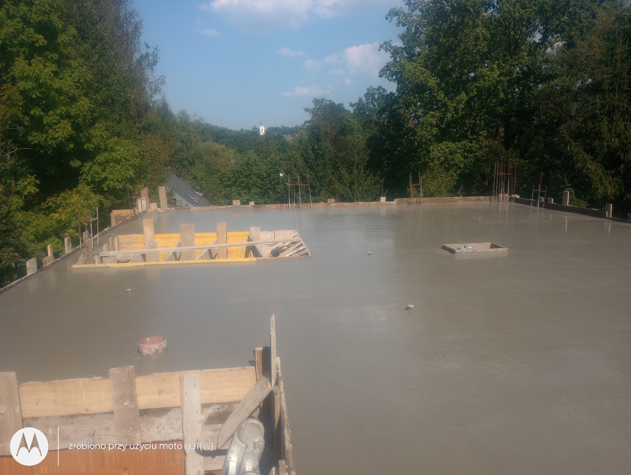 Świeżo wylany, mokry strop żelbetowy z widocznym szalunkiem, zbrojeniem i elementami instalacji, z zielonym tłem drzew i kościołem w oddali.