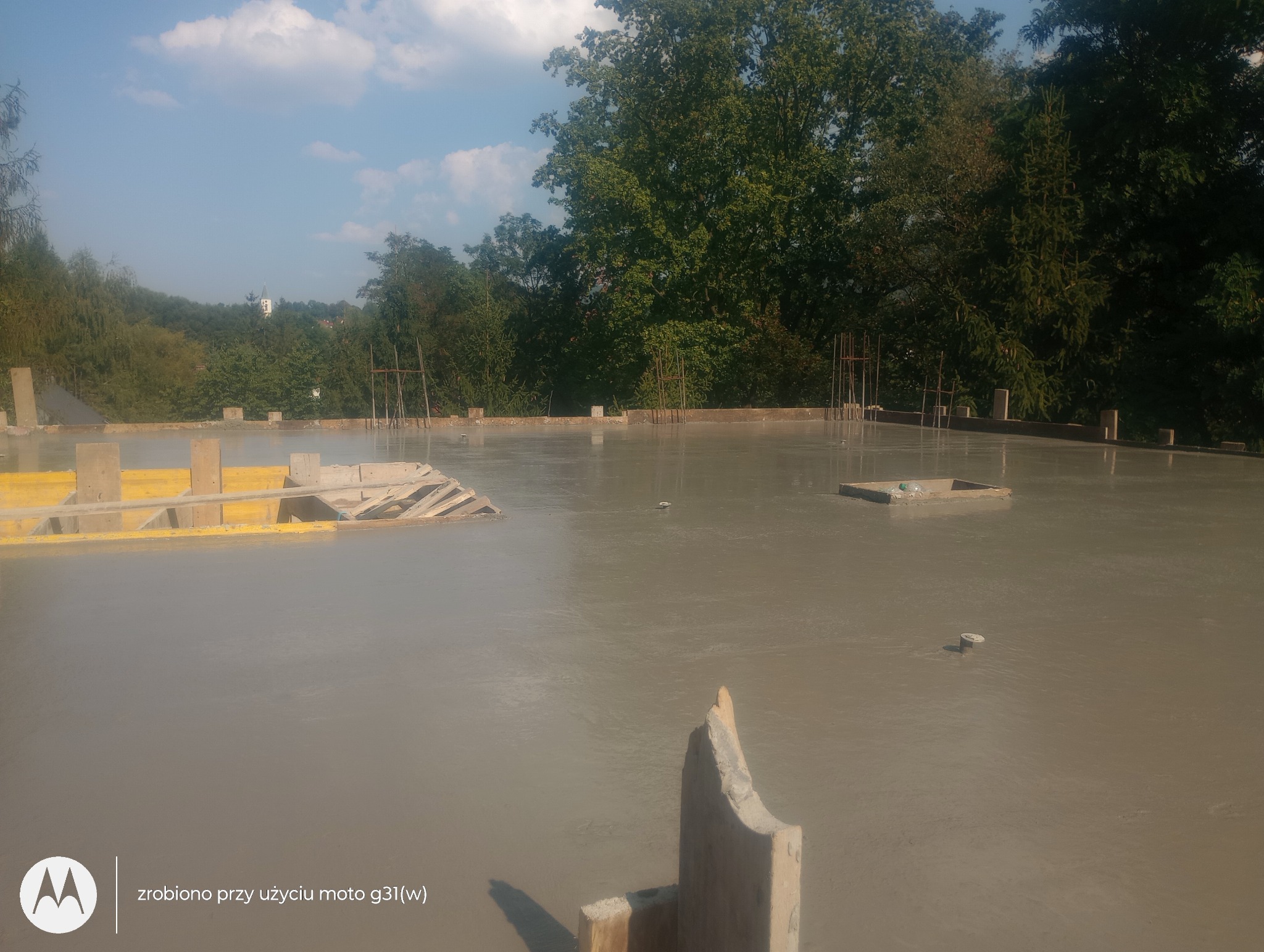 Wylany, świeży beton na stropie budynku w trakcie budowy, widoczne zbrojenia i szalunki, w tle zieleń drzew i fragment zabudowy.