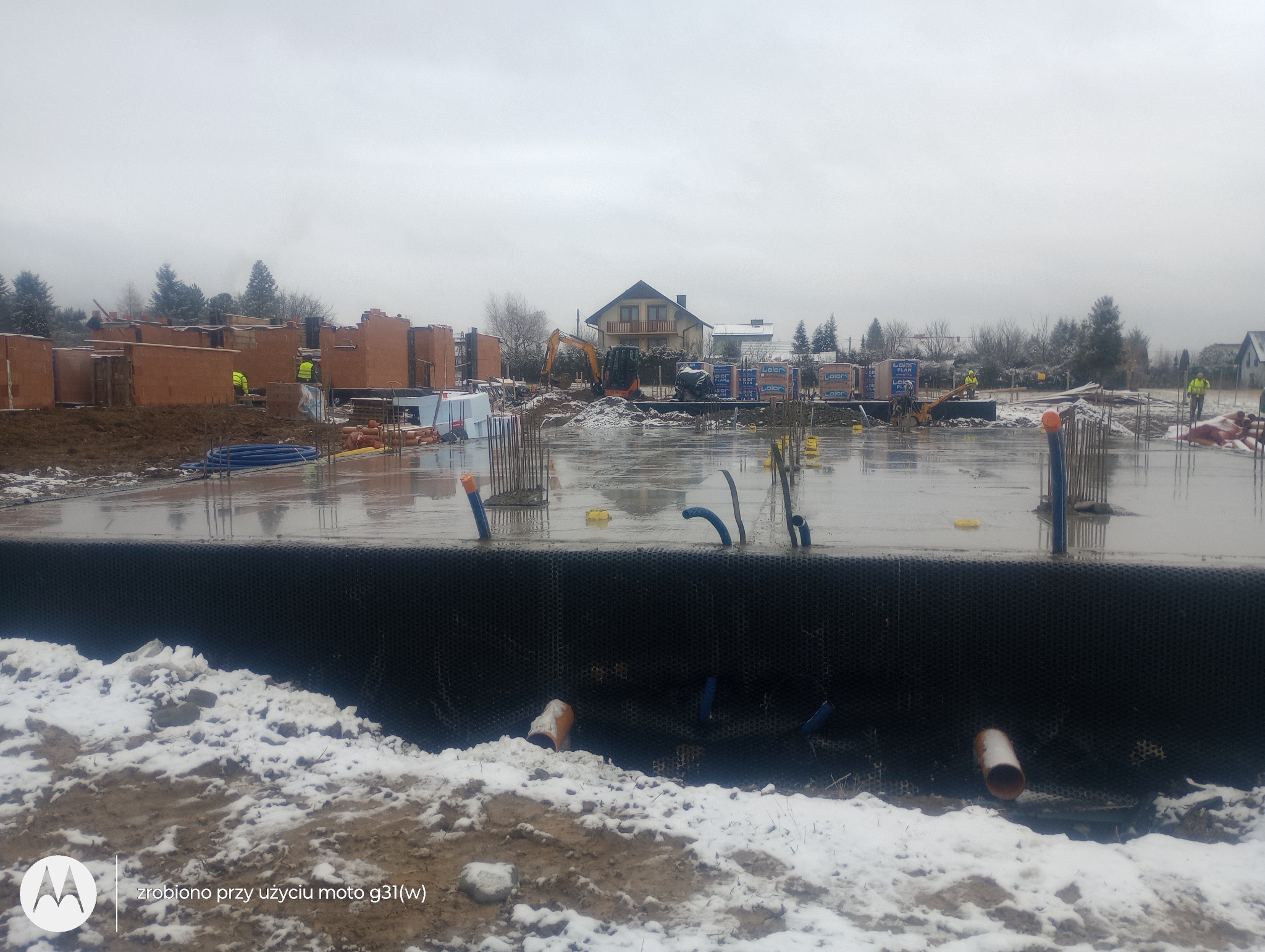 Zalana płyta fundamentowa w trakcie budowy domu jednorodzinnego, widoczne wystające pręty zbrojeniowe, rury instalacyjne, w tle częściowo wymurowane ściany z cegły, koparka i materiały budowlane...