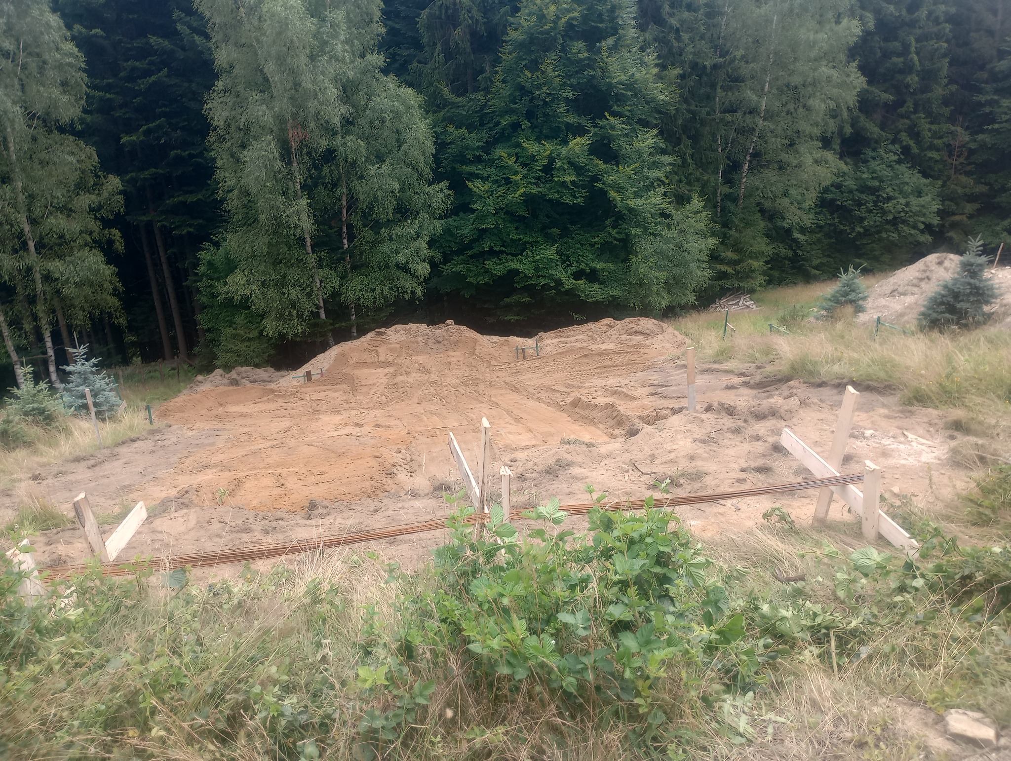 Przygotowany teren pod budowę w otoczeniu drzew, widoczne drewniane konstrukcje i pręty zbrojeniowe.