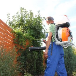 Zielony Zakątek - Mężczyzna w kombinezonie roboczym używa plecakowego opryskiwacza Stihl w ogrodzie z drewnianym płotem w tle.