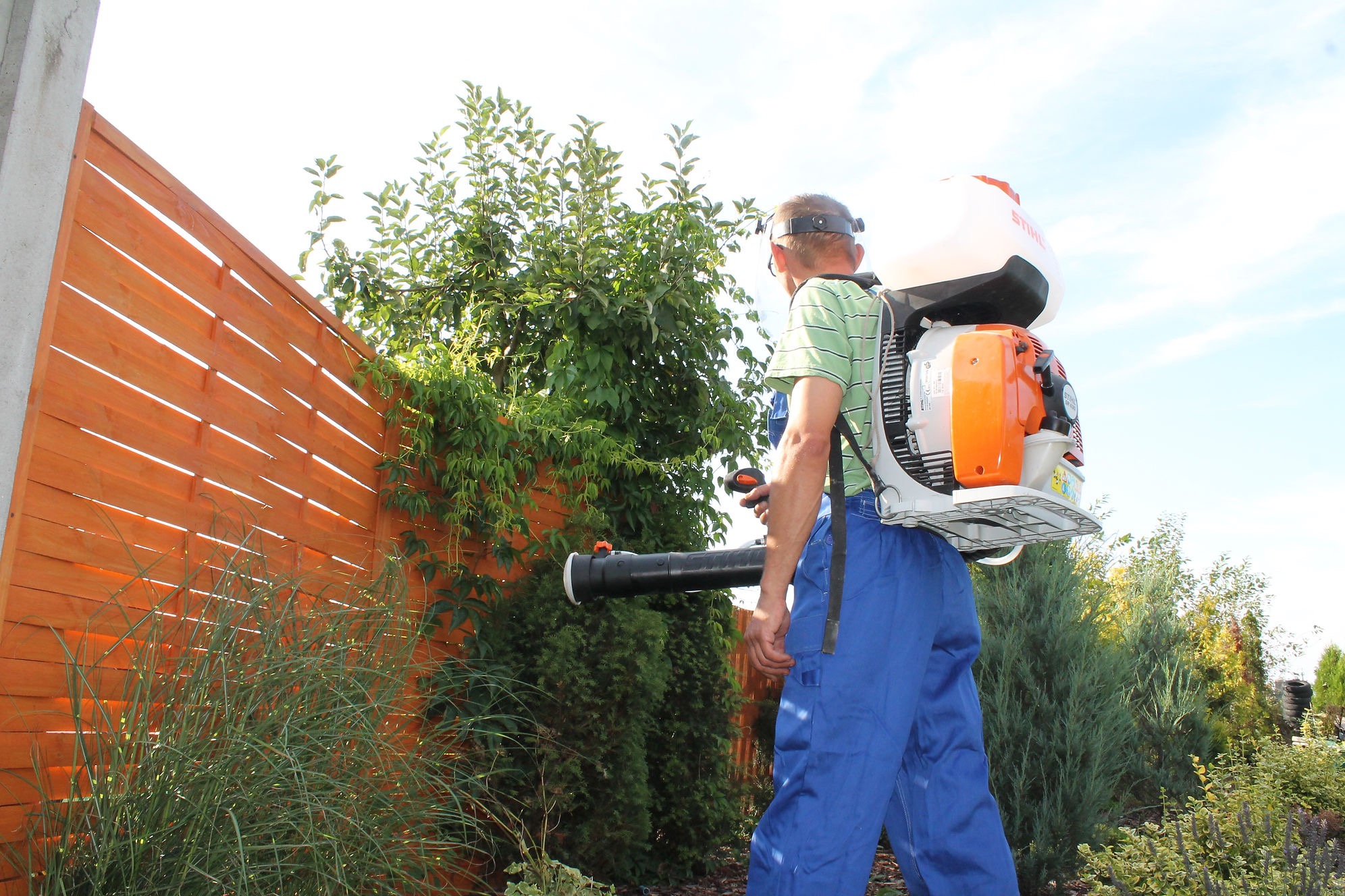 Mężczyzna w kombinezonie roboczym używa plecakowego opryskiwacza Stihl w ogrodzie z drewnianym płotem w tle.