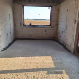 Surowy pokój z oknem na pole. Widoczne kable elektryczne w ścianach i rury ogrzewania podłogowego przed wylewką. Stan deweloperski, ściany z betonu komórkowego.