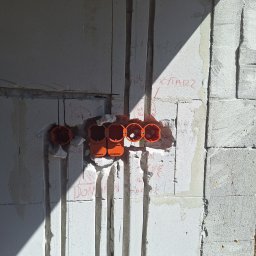 Karol Andrzejewski - Biała ściana z betonu komórkowego z wyżłobionymi bruzdami i pomarańczowymi puszkami elektrycznymi. Widoczne napisy na ścianie i cień. Instalacja elektryczna w trakcie realizacji.