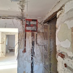 Karol Andrzejewski - Rozdzielnia elektryczna z licznymi przewodami w surowym wnętrzu podczas remontu. Widoczne ślady po kuciu w ścianach i suficie, odsłonięta instalacja elektryczna.