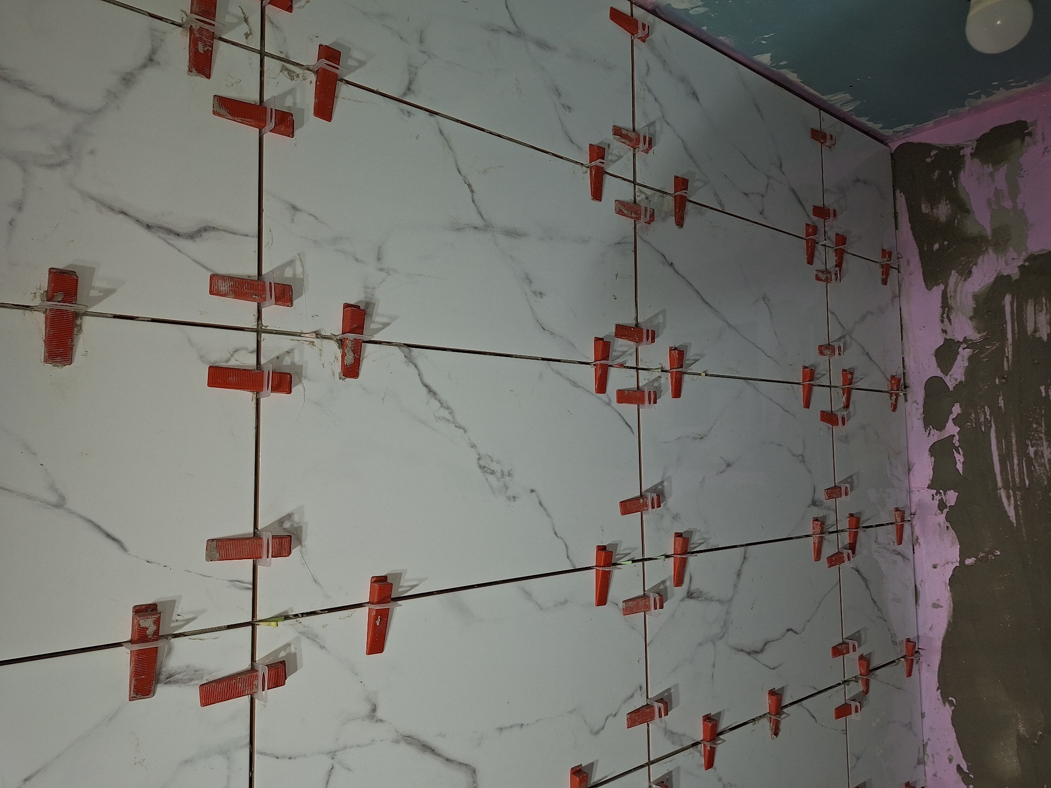 Świeżo ułożone białe płytki imitujące marmur, z czerwonymi klipsami poziomującymi i sznurkami dystansowymi, na ścianie łazienki w trakcie remontu, z widocznym surowym wykończeniem narożnika.