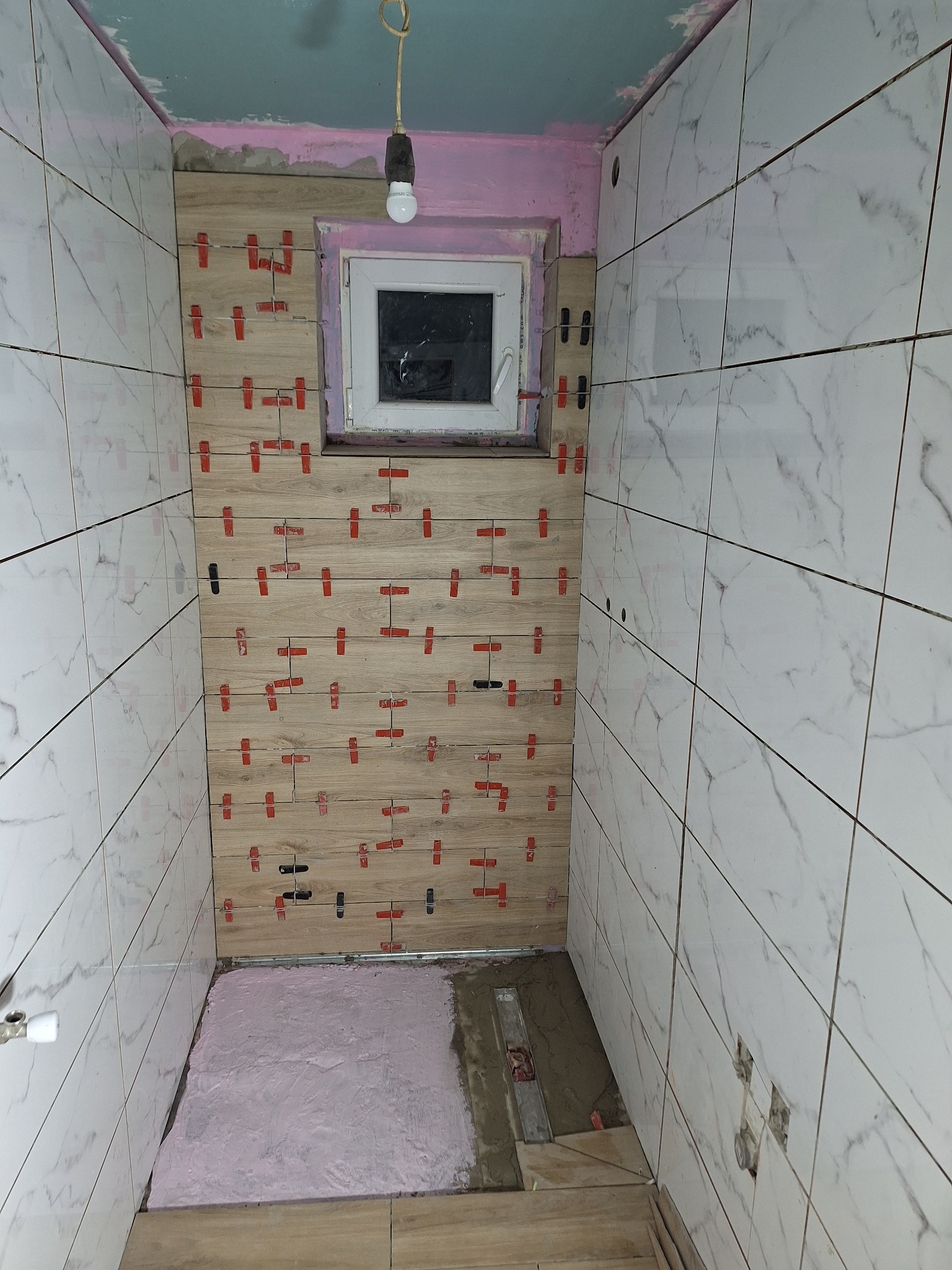 Wnętrze łazienki w trakcie układania płytek: ściany wyłożone białymi płytkami imitującymi marmur oraz drewnopodobnymi płytkami, użyte krzyżyki dystansowe, widoczny fragment wylewki podłogowej...