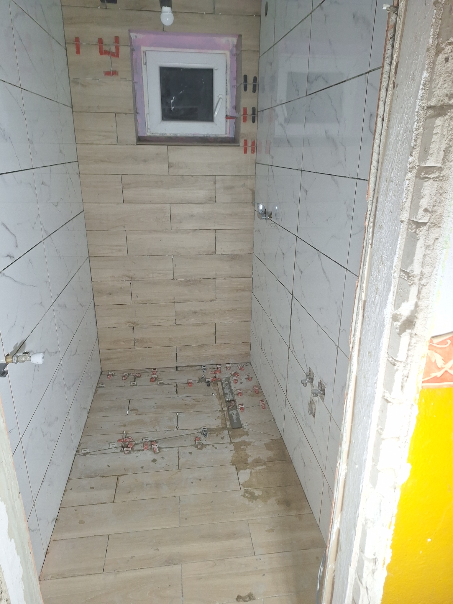 Wnętrze łazienki w trakcie układania płytek, widoczne płytki imitujące drewno na podłodze i beżowe płytki na jednej ścianie, białe marmurkowe płytki na pozostałych ścianach, plastikowe krzyżyki...