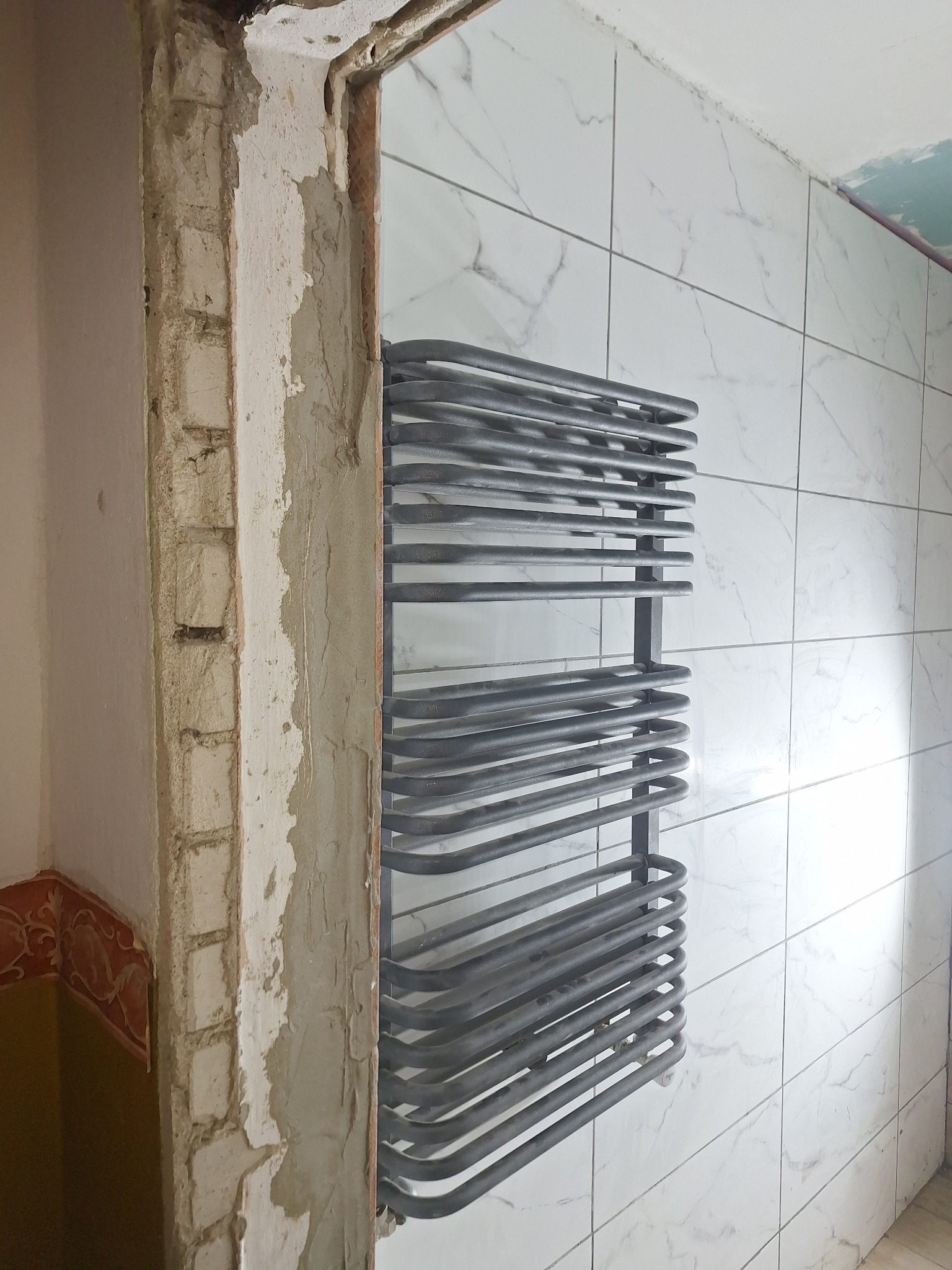 Montaż grzejnika łazienkowego drabinkowego na ścianie z białych płytek imitujących marmur, widoczne niedokończone obramowanie drzwi z surowego muru i resztkami tynku.