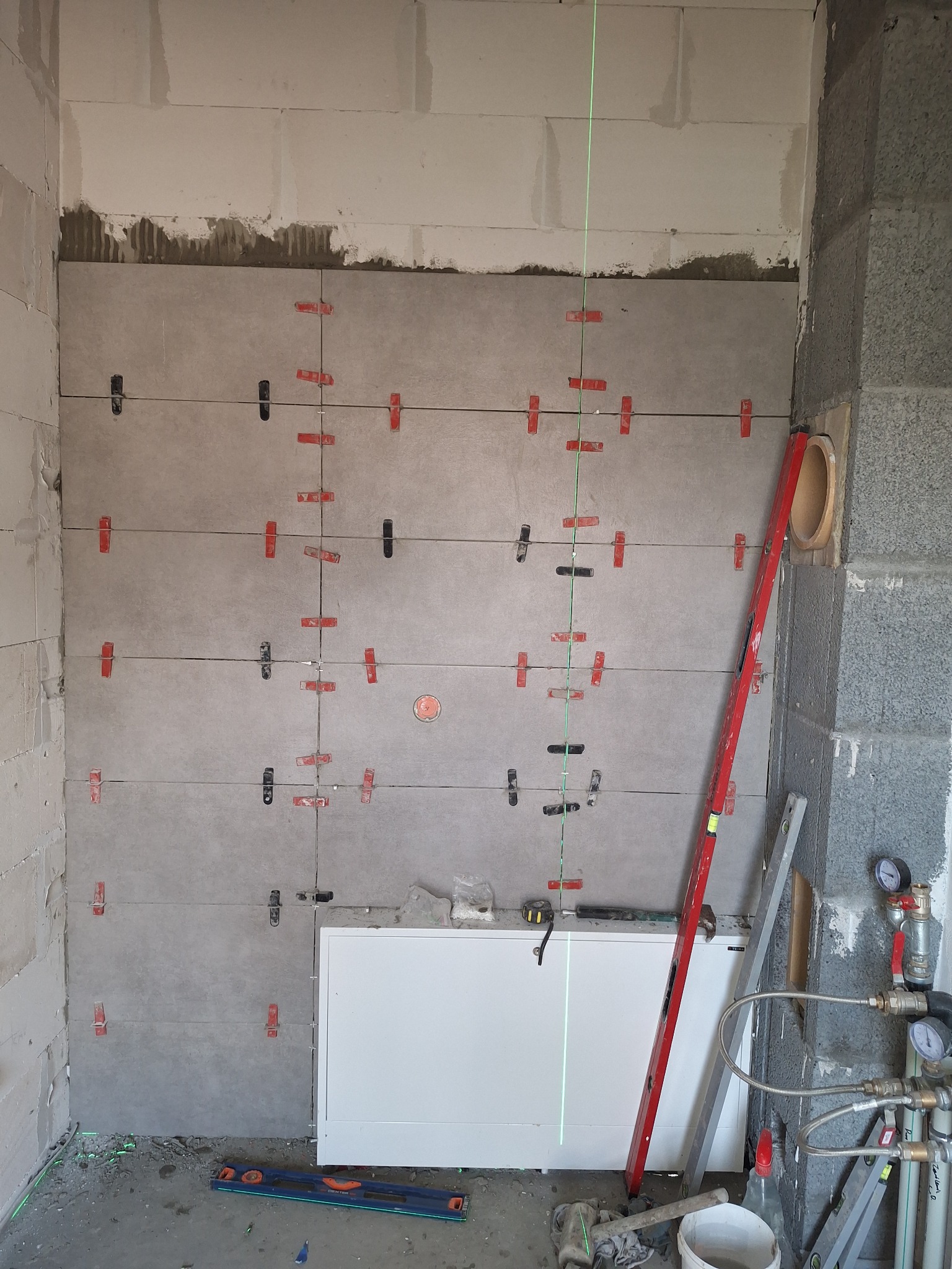 Ściana łazienki w trakcie układania szarych płytek, z użyciem poziomicy laserowej, krzyżyków dystansowych i klipsów poziomujących, widoczny fragment wanny i instalacji wodnej.