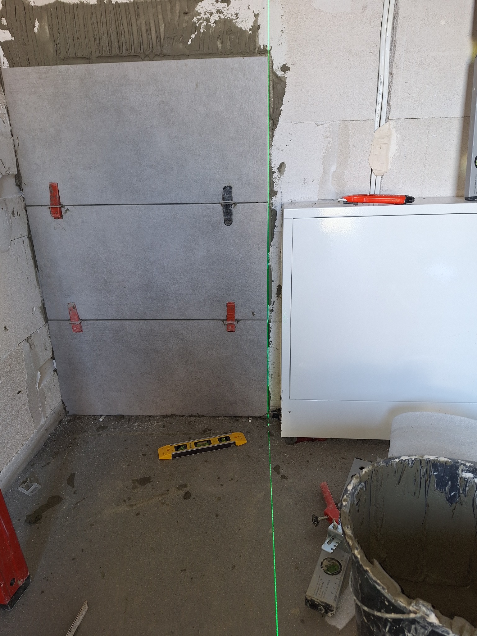 Układanie szarych płytek ściennych z użyciem poziomicy laserowej i klipsów poziomujących; widoczny fragment ściany z instalacją elektryczną i wiadro z zaprawą.