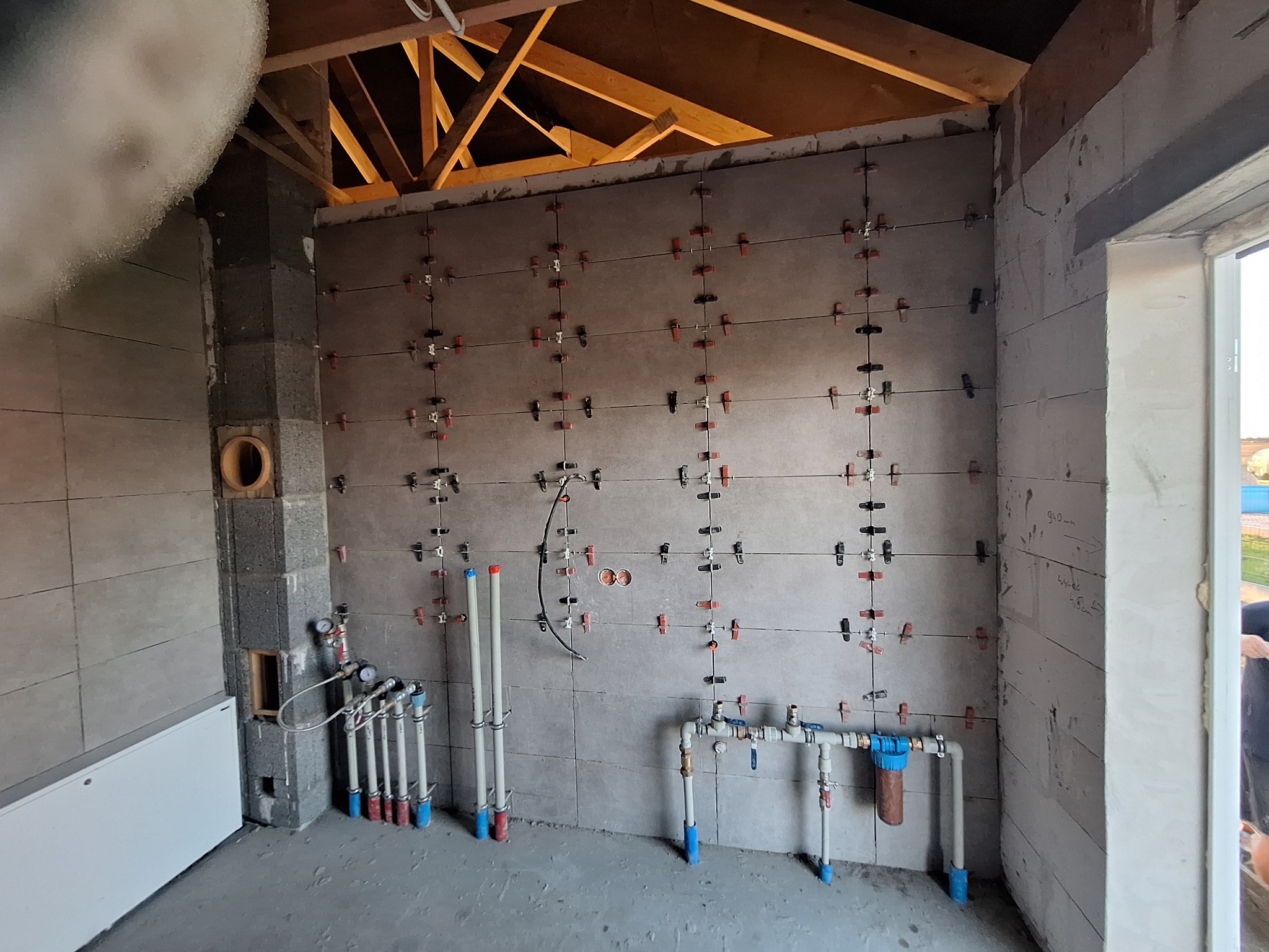 Szare płytki ścienne w trakcie układania, z widocznymi krzyżykami dystansowymi, instalacja wodna w tle, surowy stan wykończenia wnętrza.