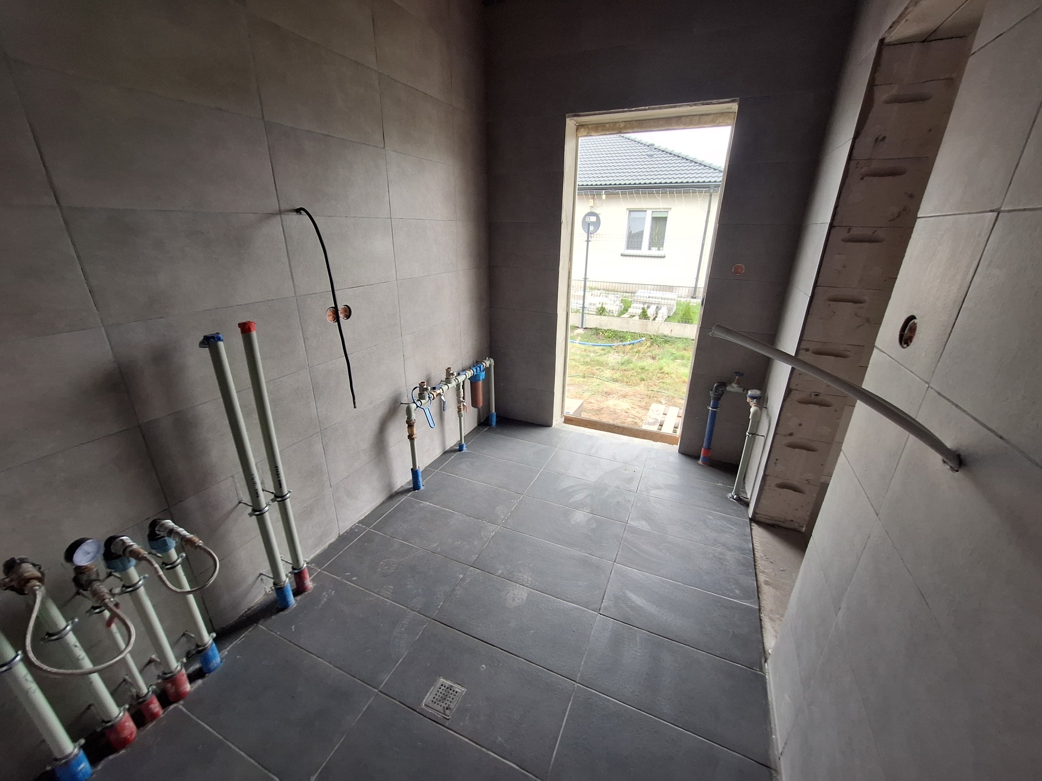 Wnętrze łazienki w trakcie prac wykończeniowych: ułożone szare płytki podłogowe i ścienne, widoczne rury instalacji wodnej z zaworami oraz fragment instalacji elektrycznej.