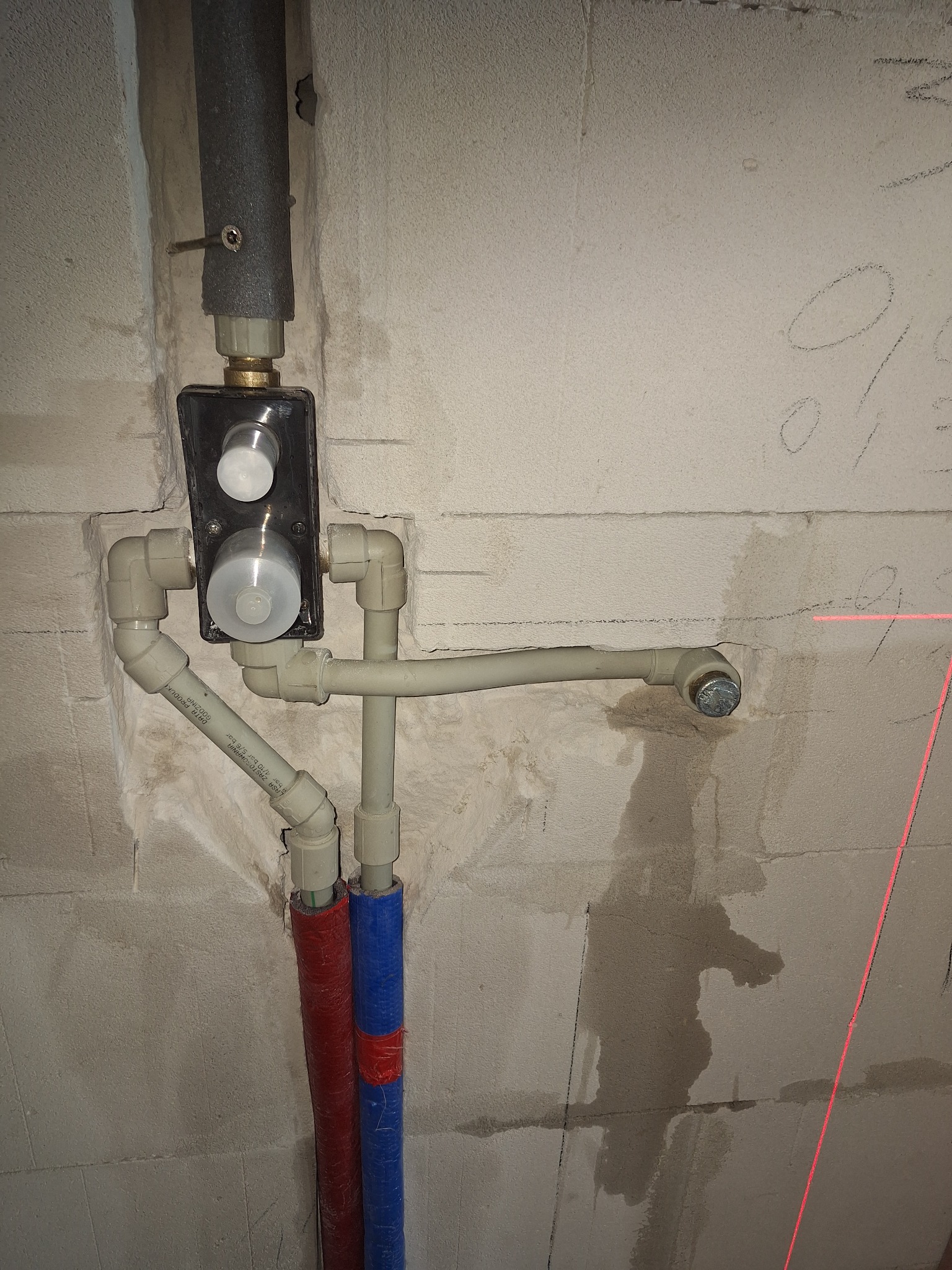 Surowy montaż baterii podtynkowej z rurami PEX w ścianie z betonu komórkowego, widoczny laserowy wskaźnik poziomu i prowizoryczne zabezpieczenie izolacji rur.