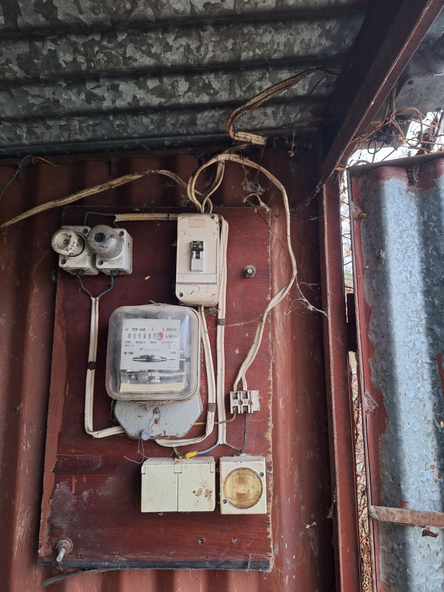 Stary licznik elektryczny z bezpiecznikami i wyłącznikiem, zamontowany na brudnej, pomalowanej na brązowo ścianie z blachy falistej, z widocznymi pajęczynami i kablami.