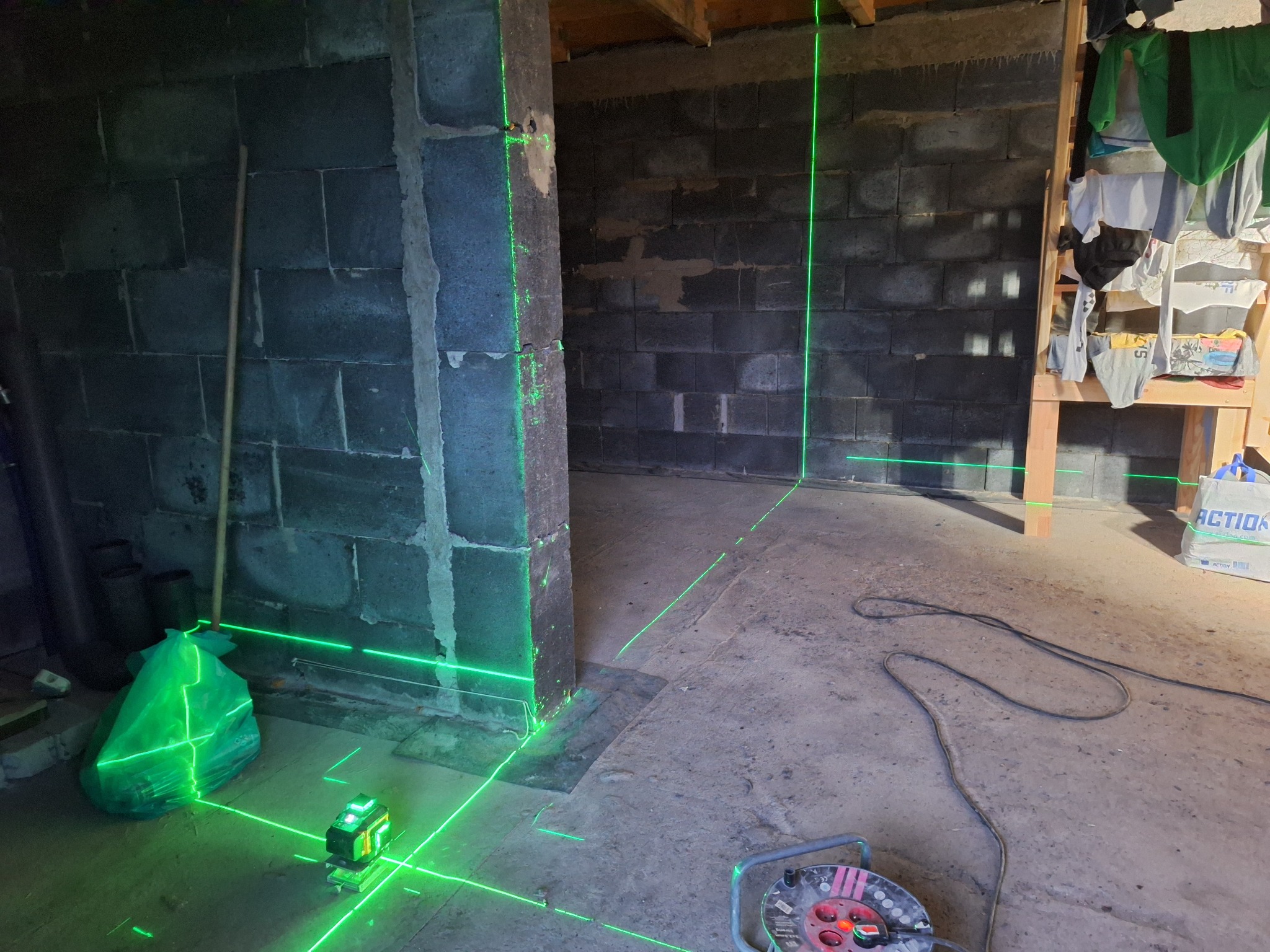 Laserowy niwelator wyznacza poziomy na surowych ścianach z bloczków betonowych w trakcie remontu piwnicy, widoczny kabel przedłużacza i worek na odpady.