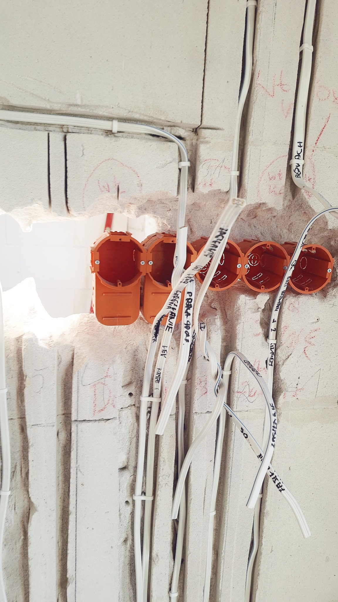 Pomarańczowe puszki elektryczne w surowej ścianie z wyprowadzonymi kablami oznaczonymi czarnym markerem. Widoczne ślady po wierceniu i kucie w betonie.