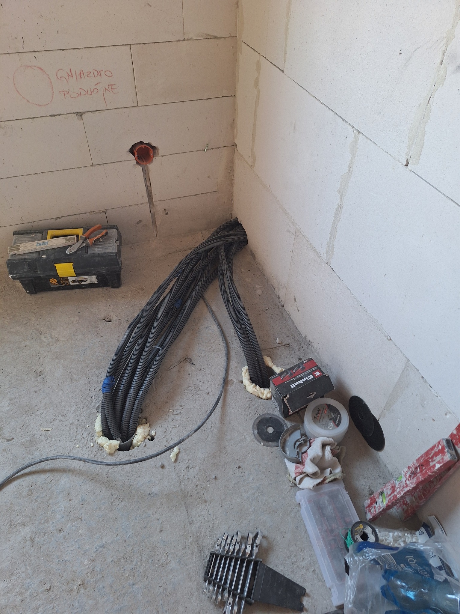 Wnętrze w trakcie instalacji elektrycznej: kable w peszlach wychodzące ze ściany, narzędzia (wiertła, tarcze) i skrzynka narzędziowa w surowym stanie.