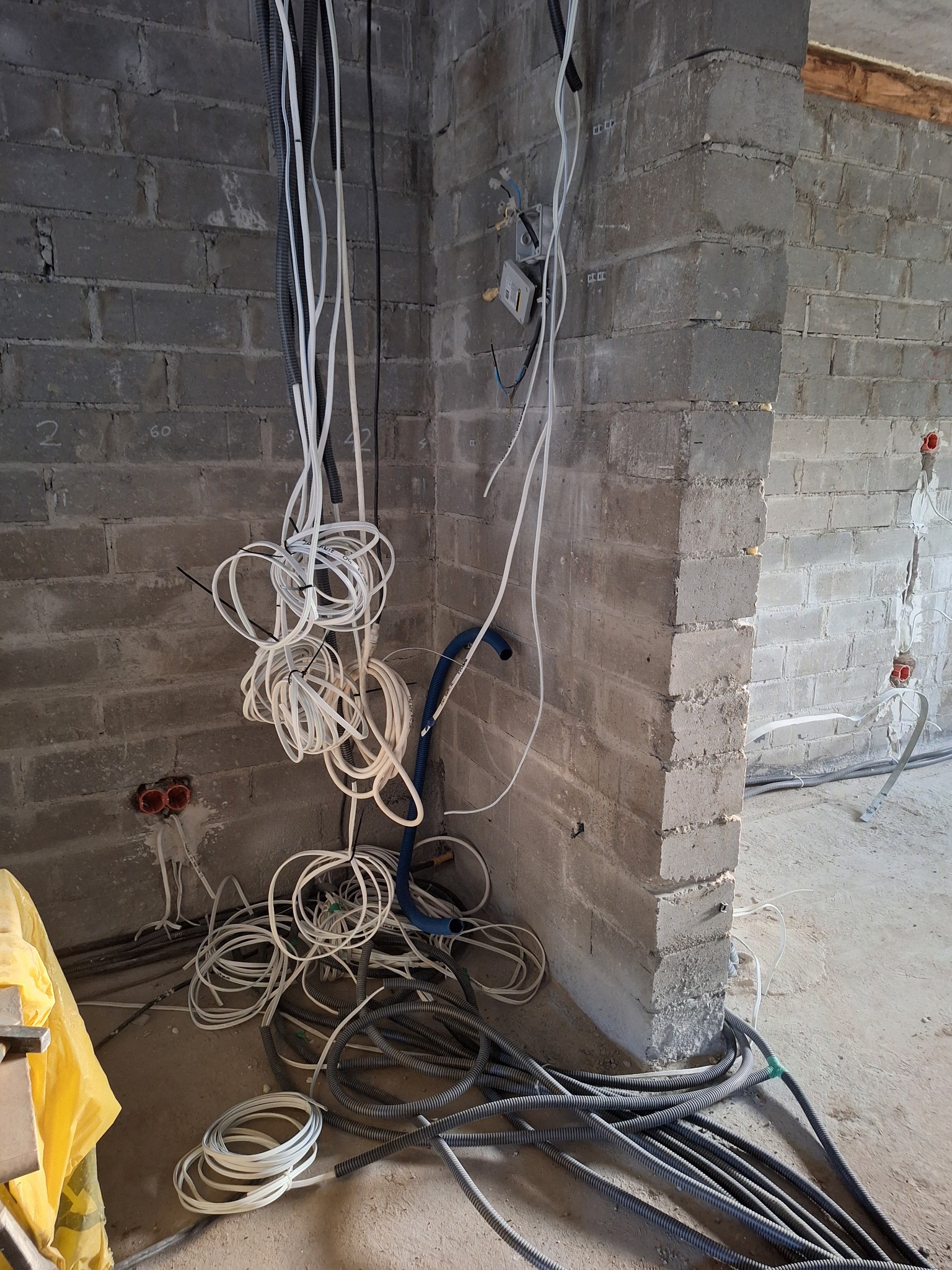 Surowy stan: przewody elektryczne i hydrauliczne zwisają ze ściany z szarej cegły w trakcie instalacji. Widoczne puszki instalacyjne i peszle na podłodze.